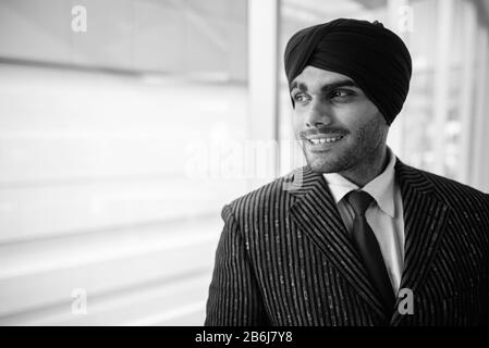 Felice giovane e bello indiano Sikh uomo d'affari che guarda attraverso la finestra Foto Stock