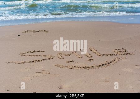 Simbolo di sorriso disegnato e sorriso di parola scritto in sabbia di spiaggia con mare blu e onde sullo sfondo Foto Stock
