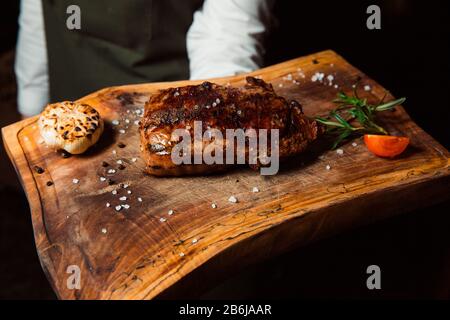 Una appetitosa bistecca di carne alla griglia preparata di fresco si trova su una tavola di legno. Cosparso di pezzi di sale, accanto ad esso si trova un rametto di razmarin e una fetta di pomodoro. Foto Stock