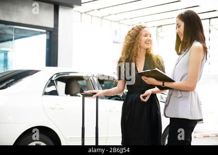 Il venditore asiatico della donna che parla e spiega circa l'assicurazione ad un cliente della donna caucasica sorridente e felice prima della consegna sopra una nuova automobile. Foto Stock