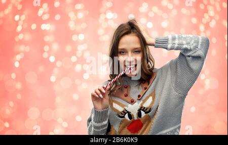 natale, persone e vacanze concetto - felice giovane donna indossando brutto maglione con modello renne mordente caramella canna sopra le luci festive sul co rosa Foto Stock