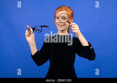 ragazza dai capelli rossi che tiene gli occhiali per la vista e una lente d'ingrandimento su uno sfondo blu Foto Stock