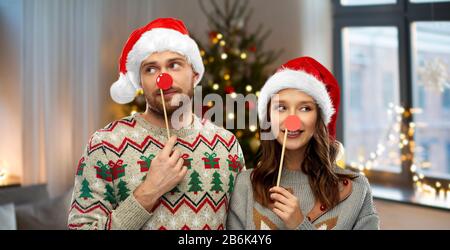 celebrazione e concetto di festa - coppia felice in ugly maglioni che posano con le punture del partito sopra le luci dell'albero di natale e lo sfondo domestico Foto Stock