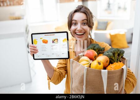 Buona donna che tiene un tablet digitale con il lancio del negozio online mentre si trova in piedi con la borsa per lo shopping piena di prodotti freschi a casa. Concetto di acquisto online tramite dispositivi mobili Foto Stock