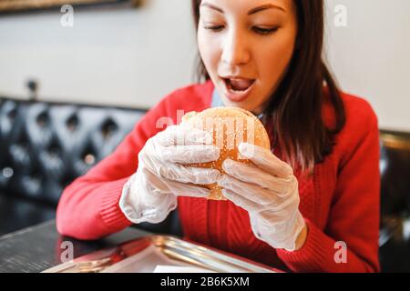 Mani di donna in guanti che tengono hamburger in moderno fast food ristorante Foto Stock