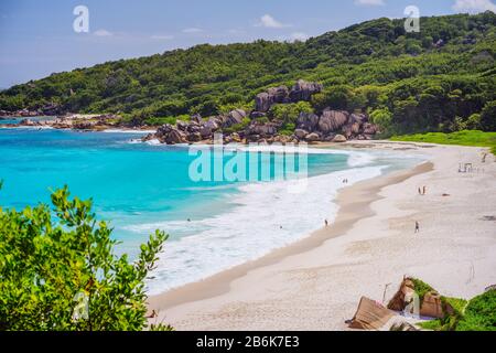 Spiaggia di Grand Anse all'isola di la Digue alle Seychelles. Lunga spiaggia di sabbia bianca con laguna blu, onde oceaniche e massi di granito sullo sfondo. Foto Stock