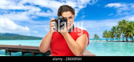 concetto di viaggio, turismo e vacanza - felice donna fotografo in abito rosso con fotocamera digitale su sfondo tropicale spiaggia in polinesia francese Foto Stock