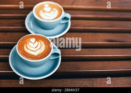 Due tazze di cappuccino con latte art su sfondo di legno. Bella schiuma, coppe in ceramica blu, composizione diagonale, luogo per il testo. Foto Stock