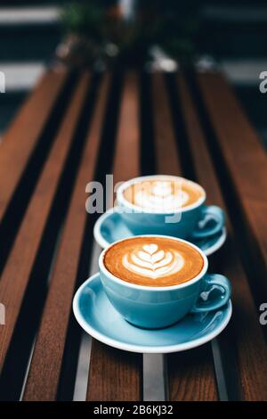 Due tazze di cappuccino con latte art su sfondo di legno. Bella schiuma, coppe in ceramica blu, composizione diagonale, luogo per il testo. Foto Stock