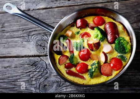 frittata al forno o frittata con salsicce, broccoli, pomodori ciliegini, formaggio fuso in una padella su un tavolo rustico in legno, vista orizzontale dall'alto, cl Foto Stock