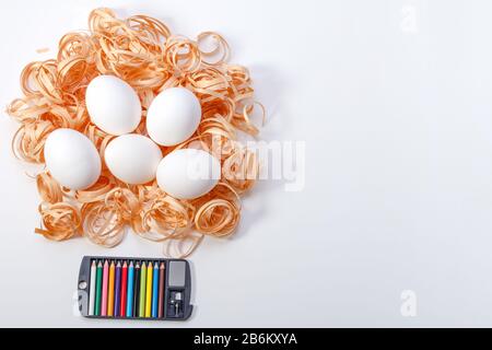 Cinque uova bianche disposte su un nido di trucioli di legno con una selezione di matite colorate in una scatola nera su uno sfondo bianco, pronte per essere colorate per Ea Foto Stock