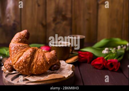Croissant e tulipani rossi. Colazione di Pasqua con espresso fresco aromatico. Caffè con dolci, fiori su un tavolo di legno. Spazio di copia. Foto Stock