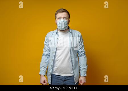 Uomo in una maschera medica protettiva, è molto impaurito di contrarre il virus, pugni radicati e guarda in camera in sgomento. Foto Stock