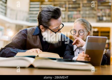 Bella ragazza intelligente in occhiali e vecchio insegnante di uomo che ha una lezione e discussione in libreria vintage. La ragazza tiene il lettore digitale del ebook o del tablet e. Foto Stock