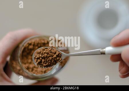 Bicchiere di vetro con caffè in mano. Una mano tiene un cucchiaio pieno di caffè istantaneo. Mucchio di chicchi di caffè istantanei in cucchiaio di metallo sul vaso di vetro. Caffeina dr Foto Stock