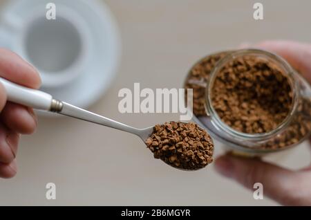 Bicchiere di vetro con caffè in mano. Una mano tiene un cucchiaio pieno di caffè istantaneo. Mucchio di chicchi di caffè istantanei in cucchiaio di metallo sul vaso di vetro. Caffeina dr Foto Stock
