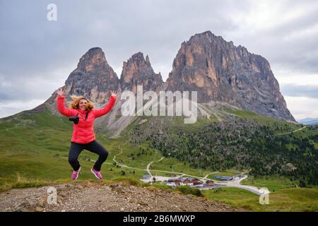 Felice fotografo saltando con le braccia in alto in aria, indossando una giacca rossa luminosa, di fronte al monte Sassolungo (Saslonch, Langkofel), Dolomiti, Foto Stock