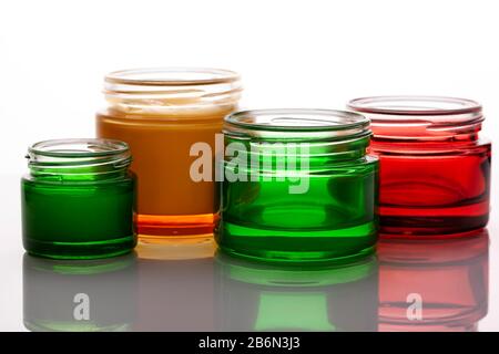 Barattoli colorati in vetro per cosmetici vuoti e pieni di crema su sfondo luminoso. Riflessione su una superficie lucida. Luce di sfondo Foto Stock