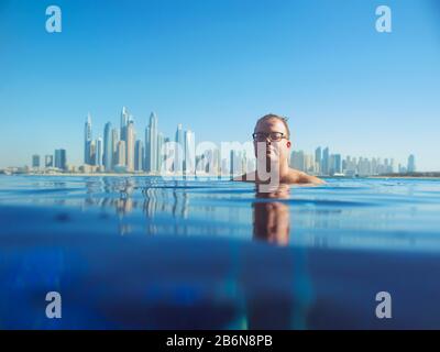 Ritratto di allegro e rilassato uomo rosso caucasico europeo in occhiali in piscina sulla Dubai moderna (Emirati Arabi Uniti) e sullo sfondo del mare Foto Stock