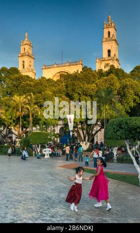 Ragazze giovani a Plaza Grande, Catedral de San Ildefonso dietro alberi di alloro in lontananza, al tramonto, Merida, Stato dello Yucatan, Messico Foto Stock