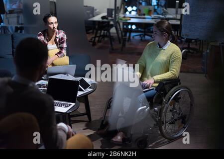 Giovane donna portatrice di handicap con colleghi che lavorano in ufficio. Sorride e si appassiona al flusso di lavoro. Esecuzione nello spazio di co-working. Office pe Foto Stock