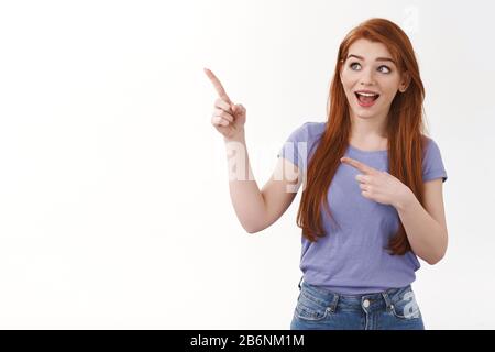 Curioso gioioso sorridente donna rossa in t-shirt viola, in piedi intrigato o intrigato, puntando guardando angolo superiore sinistro divertito, vedere interessante Foto Stock