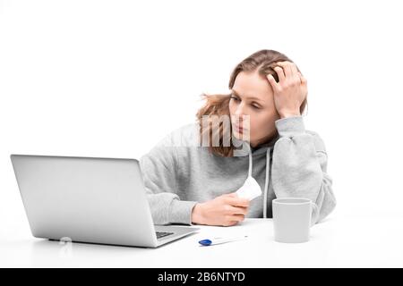 Giovane donna con cappuccio seduto al tavolo e tenendo tovagliolo mentre legge sui sintomi su internet utilizzando il computer portatile Foto Stock