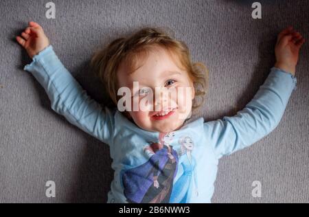 Cheeky ritratto di 3 anni ragazza avendo divertimento a casa guardando in su sorridendo alla macchina fotografica Foto Stock