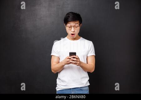 Tecnologia, gadget e concetto di persone. Stupito e impressionato il bel ragazzo asiatico reagire alle notizie emozionanti che ha letto in linea, tenendo il telefono mobile, stare Foto Stock
