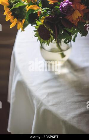 Un bouquet con rustici fiori selvatici estivi, girasoli è sul tavolo con una tovaglia bianca. Stile di vita. Spazio di copia. Film tinting. Moody foto. Foto Stock