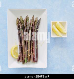 Asparagi freschi organici cotti al vapore con fettine di limone su porcellana bianca su fondo blu in composizione piatta. Foto Stock