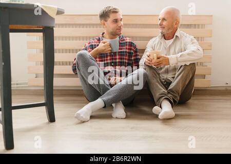 Un ritratto di adulto hipster figlio e padre senior seduto sul pavimento all'interno di home. Foto Stock