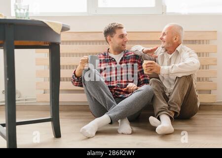 Un ritratto di adulto hipster figlio e padre senior seduto sul pavimento all'interno di home. Foto Stock