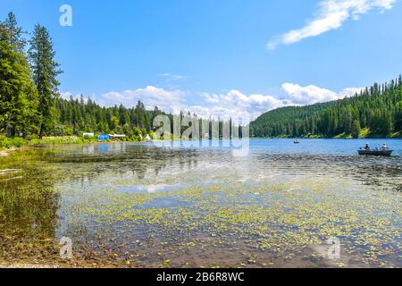 Due persone pescano da una piccola barca a remi sul lago Granite con case rurali sul lungomare e una teepee nelle panoramiche montagne dell'Idaho settentrionale degli Stati Uniti Foto Stock