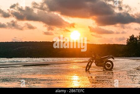 Una moto d'argento si trova in spiaggia al tramonto Foto Stock