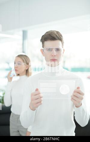 Ritratto di giovane ingegnere software in occhiali intelligenti che analizza le statistiche su un innovativo tablet in ufficio Foto Stock