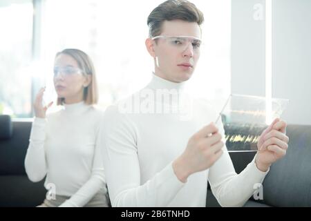 Ritratto di un ragazzo serio e moderno in occhiali intelligenti che registra la voce su un dispositivo portatile trasparente e che confronta le tracce audio Foto Stock