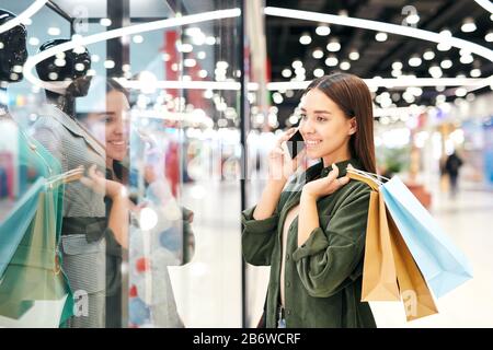 Ragazza studentesca sorridente con borse di carta in piedi alla vetrina e guardando vestito mentre chiedendo consiglio di amico per telefono Foto Stock