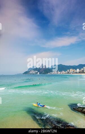 Un surfista maschio che entra nell'Oceano Atlantico alle rocce Arpoador con spiaggia Ipanema, quartiere e le montagne Dois Irmaos sullo sfondo Foto Stock