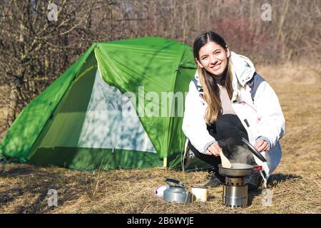 ragazza allegra campeggio da sola cucina di fronte alla sua tenda. tempo per l'aria aperta Foto Stock