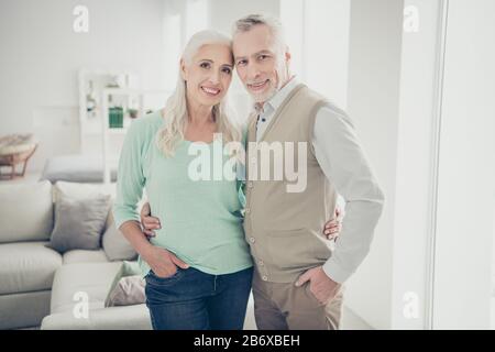 Portrait dei nonni attraenti cudling che osservano i jeans vestiti jeans denim pantaloni turchese marrone pullovers che stanno in grande stanza Foto Stock