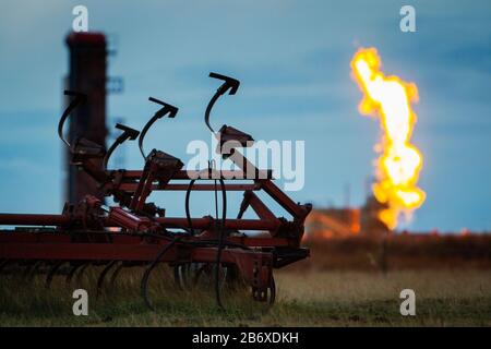 Un vecchio timone si siede in primo piano mentre le fiamme dal fuoco di metano e gas da un pozzo di petrolio si illumina sullo sfondo. Il pozzo petrolifero è uno dei molti pozzi di produzione proprio fuori Williston nel North Dakota. L'area fa parte del settore petrolifero di Bakken, dove la tecnologia del fracking lo ha reso redditizio finché il prezzo del petrolio rimane al di sopra dei 46 dollari al barile. Foto Stock