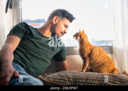primo piano di giovane uomo e gatto tabby guardando profondamente l'un l'altro di fronte ad una finestra Foto Stock
