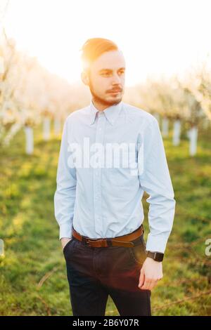 Ritratto all'aperto di elegante uomo di sposo barbuto in camicia chiara e pantaloni scuri. Bell'uomo sorridente all'aperto, camminando nel giardino primaverile con fioritura Foto Stock