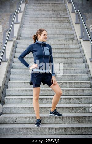 Giovani donne sportive vestite di abbigliamento sportivo davanti alle scale. Sportliche junge Frau mit Sportbekleidung steht vor einer Treppe. Foto Stock