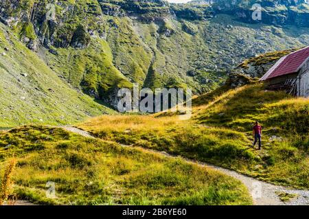 Uomo che cammina su un sentiero di montagna vicino chalet di montagna con la montagna sullo sfondo Foto Stock