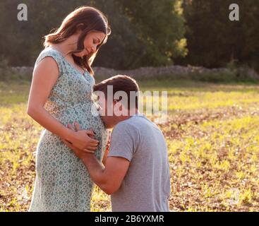 Giovane donna incinta in piedi in un campo, con la sua partner inginocchiata carezzandola sul pancino Foto Stock