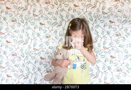 Una bambina carina con un orso di teddy starnutisce in un tovkin perché ha ottenuto un'infezione virale. Un bambino malato con tosse e starnuti al naso che cola. Aller Foto Stock