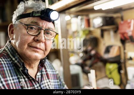 Ritratto artigiano con torcia a LED con lampada frontale seduta in workhop con attrezzi e attrezzature sullo sfondo. Vecchio insegnante grigio maturo del handyman con Foto Stock