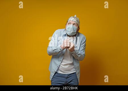 L'uomo di oddball in una maschera medica è molto impaurito di contrarre il virus, ha messo su un cappello di foglio di stagno e guarda al piano superiore in sgomento. Foto Stock